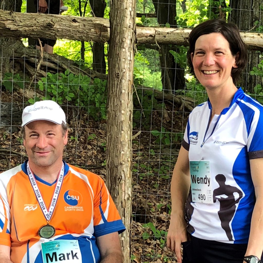 Two people in the woods smile while wearing athletic clothing and bibs